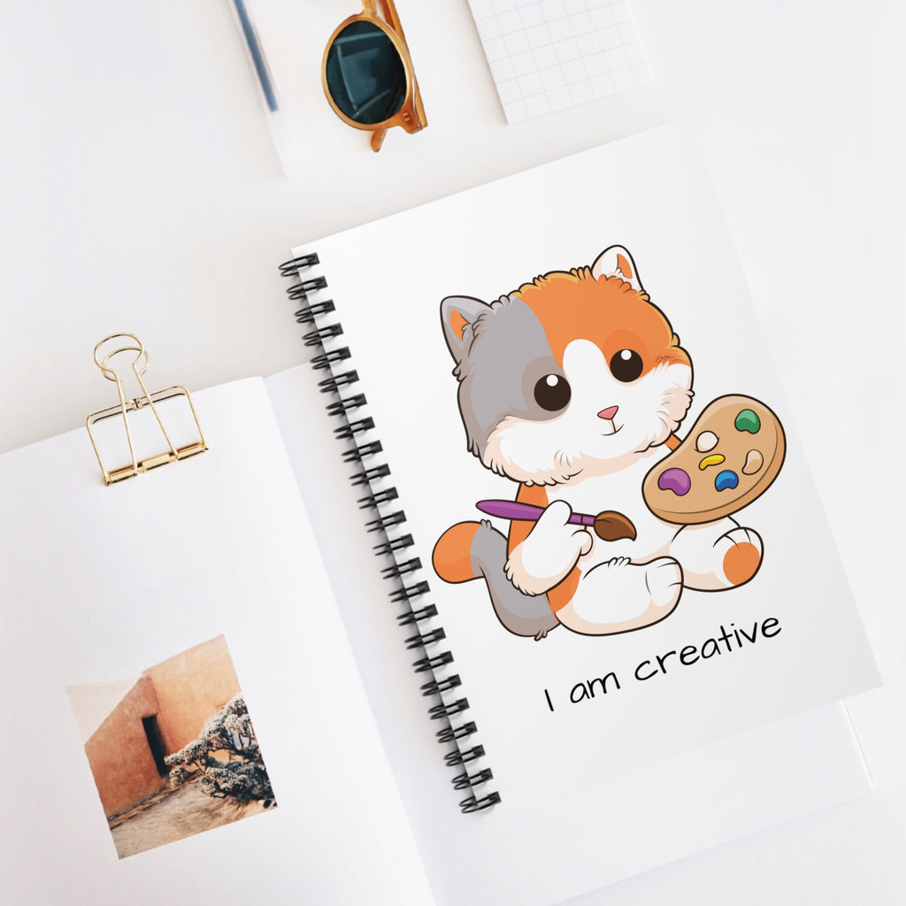 White spiral notebook with a picture of a cat that says I am creative. The notebook is laying closed on a desk.