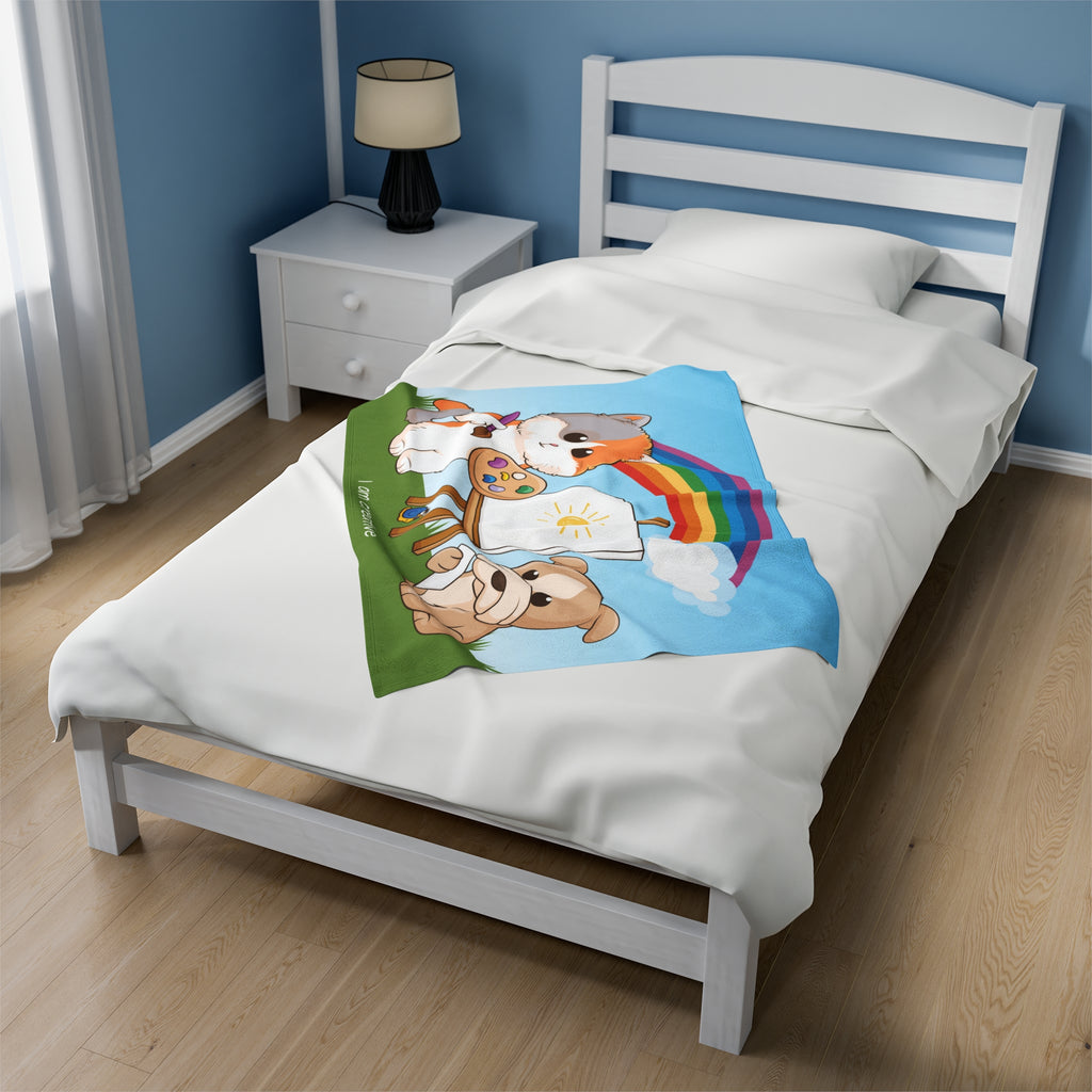 A 30 by 40 inch blanket on a twin-sized bed in a bedroom. The blanket has a scene of a cat painting on a canvas next to a dog, a rainbow in the background, and the phrase "I am creative" along the bottom.