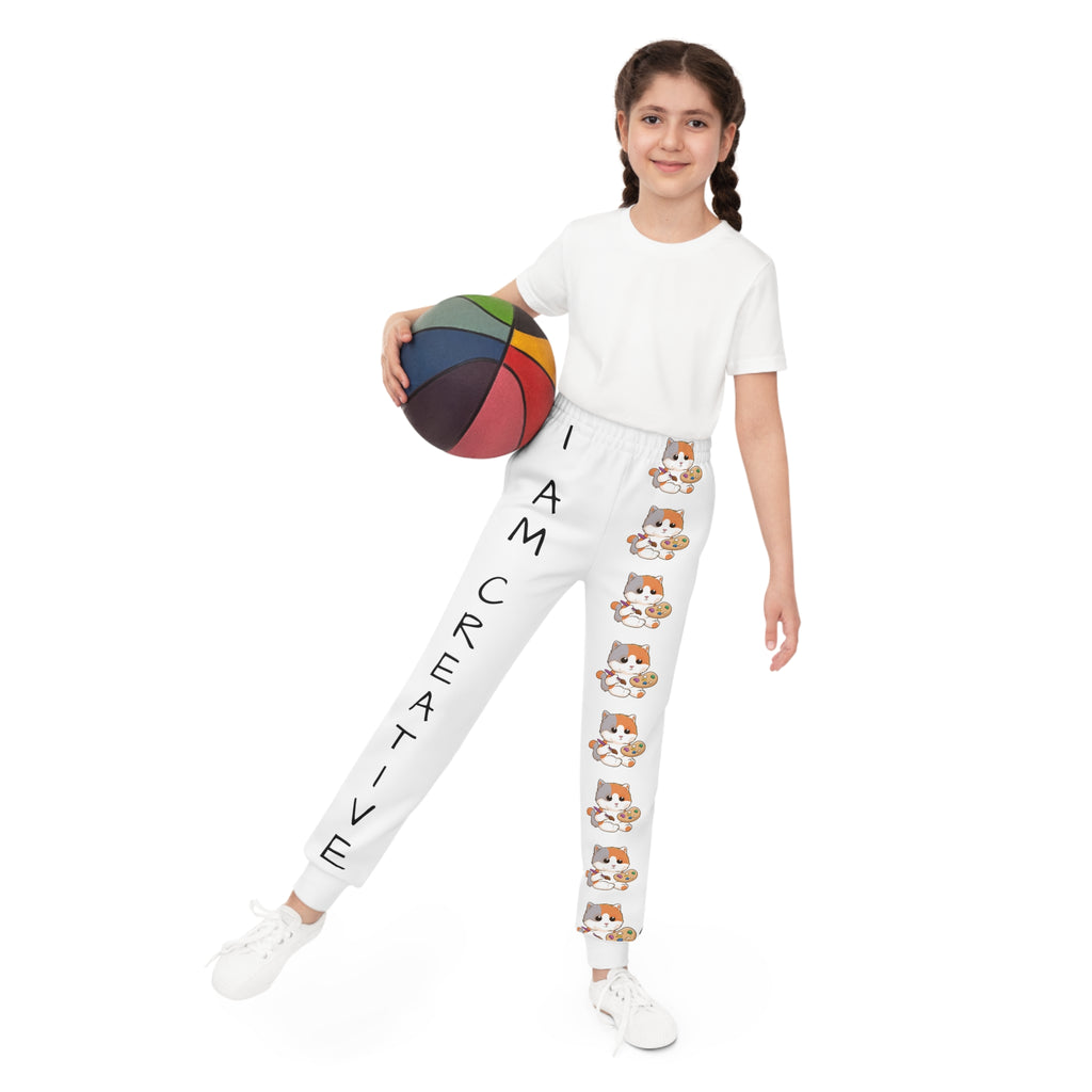 Front-view of a girl holding a basketball and wearing white sweatpants. The pants have a line of cats down the front left leg and the phrase "I am creative" down the front right leg.