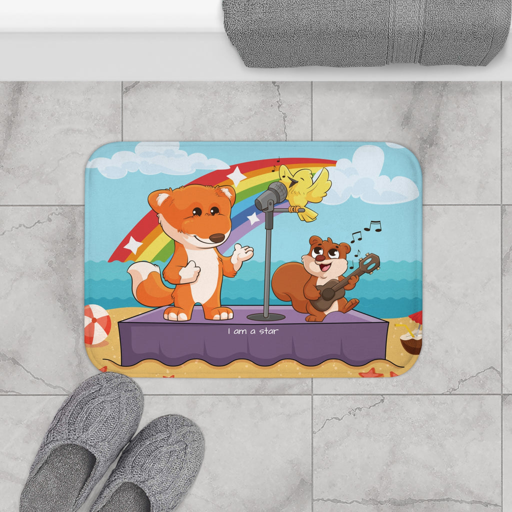 A 24 by 17 inch bath mat on the tiled floor of a bathroom. The bath mat has a scene of a fox singing with a bird and squirrel on a stage on the beach with a rainbow in the background and the phrase "I am a star" along the bottom.