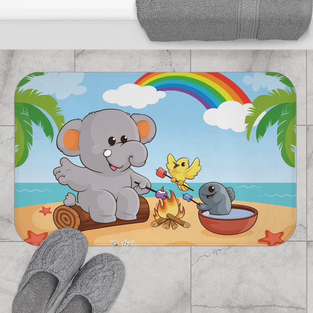 A 34 by 21 inch bath mat on the tiled floor of a bathroom. The bath mat has a scene of an elephant having a bonfire with a bird and fish on the beach, a rainbow in the background, and the phrase "I am calm" along the bottom.