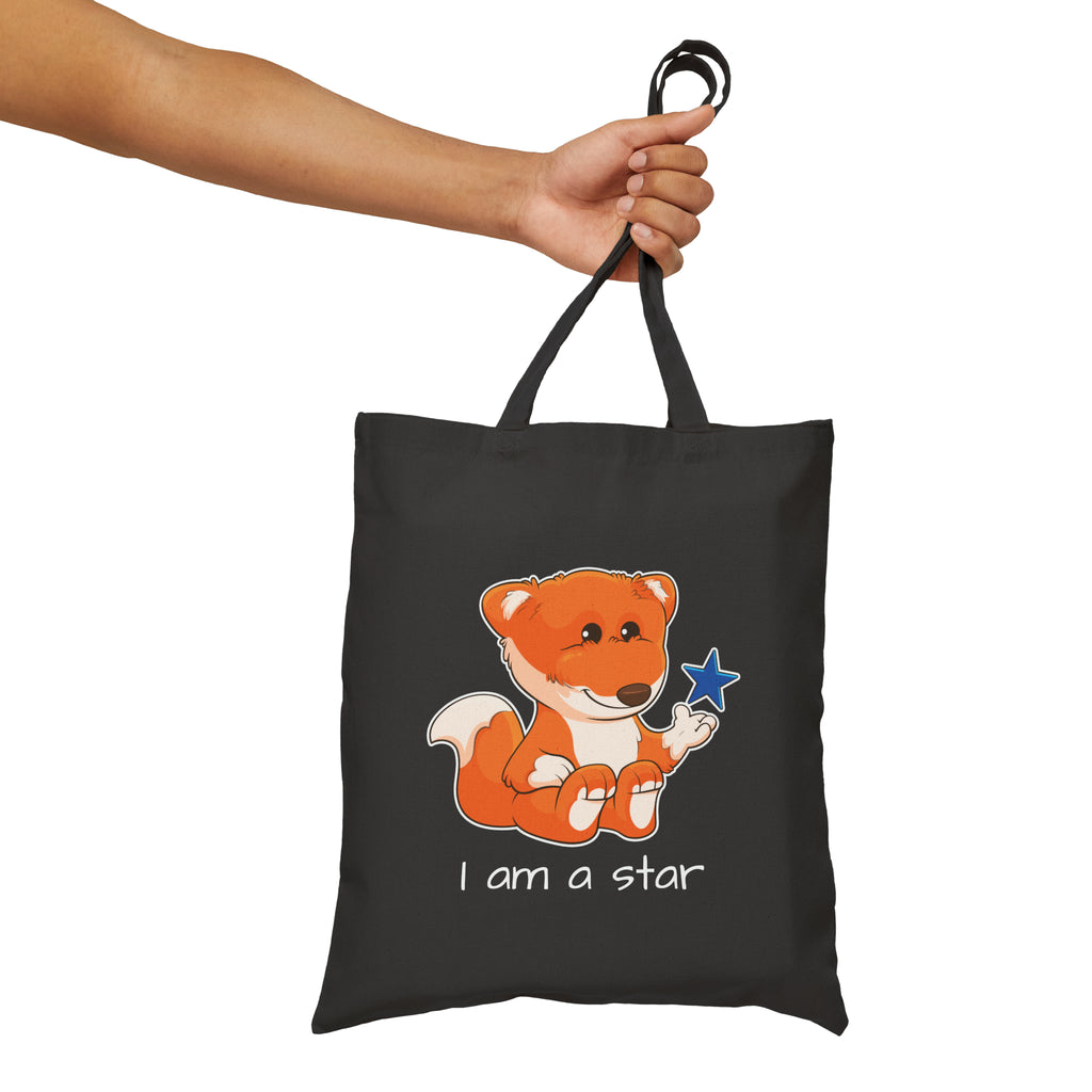 A hand holding a black tote bag with a picture of a fox that says I am a star.