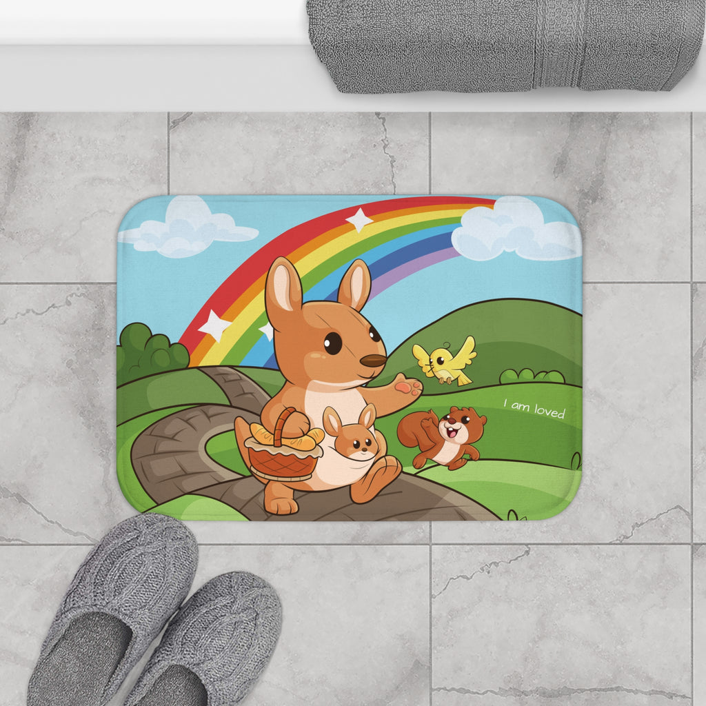 A 24 by 17 inch bath mat on the tiled floor of a bathroom. The bath mat has a scene of a kangaroo walking on a path through rolling hills with a rainbow in the background and the phrase "I am loved".
