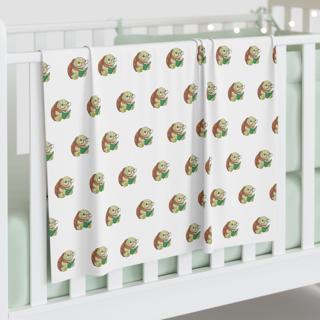 A white swaddle blanket with a repeating pattern of a turtle. The blanket is draped over the side of a baby crib.