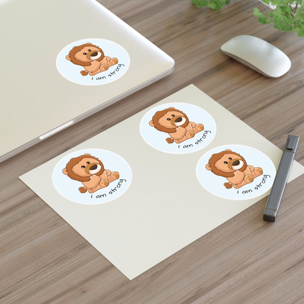 A sheet of 3 round stickers with a picture of a lion that says I am strong. The sticker sheet sits on a table next to a laptop with the fourth sticker on it.