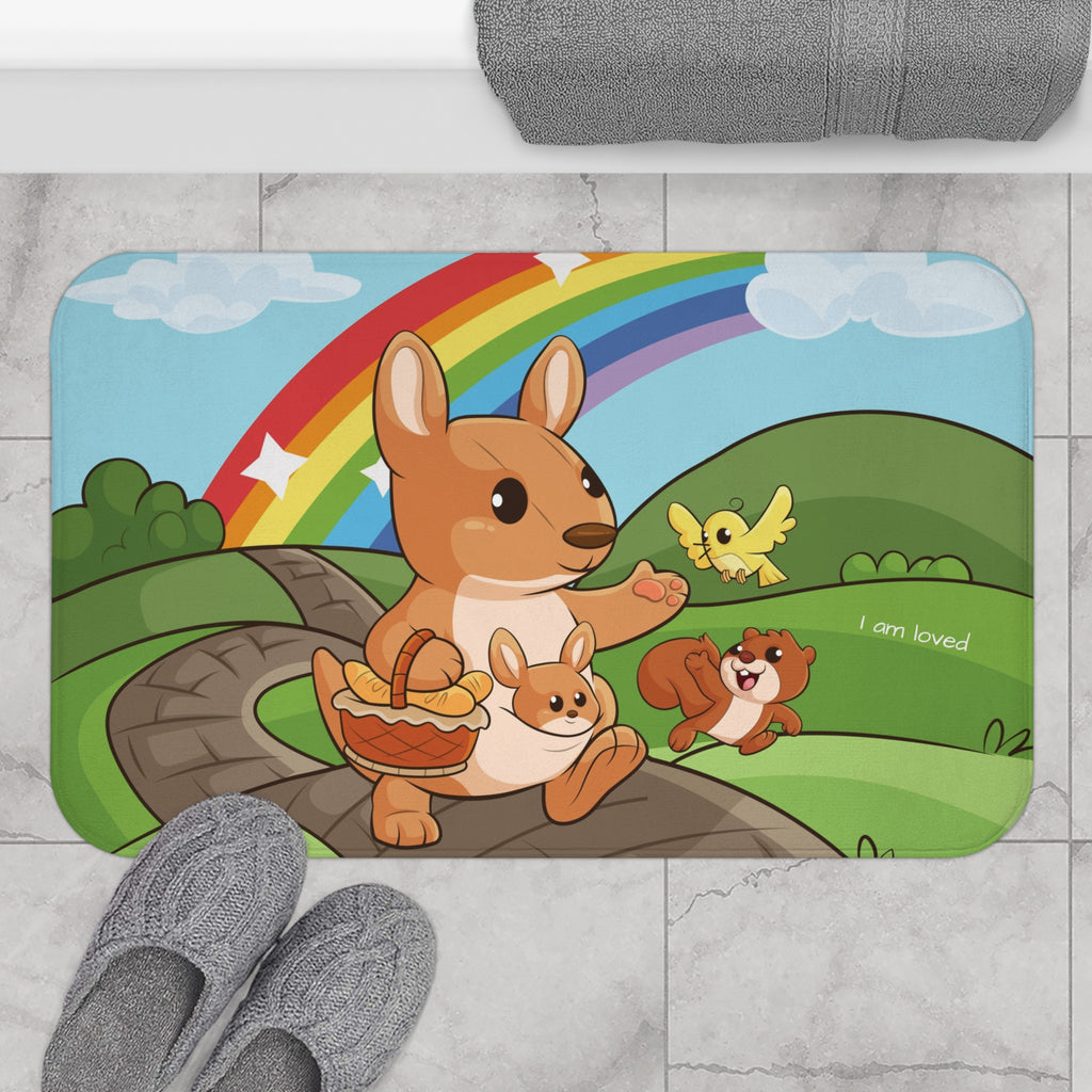 A 34 by 21 inch bath mat on the tiled floor of a bathroom. The bath mat has a scene of a kangaroo walking on a path through rolling hills with a rainbow in the background and the phrase "I am loved".