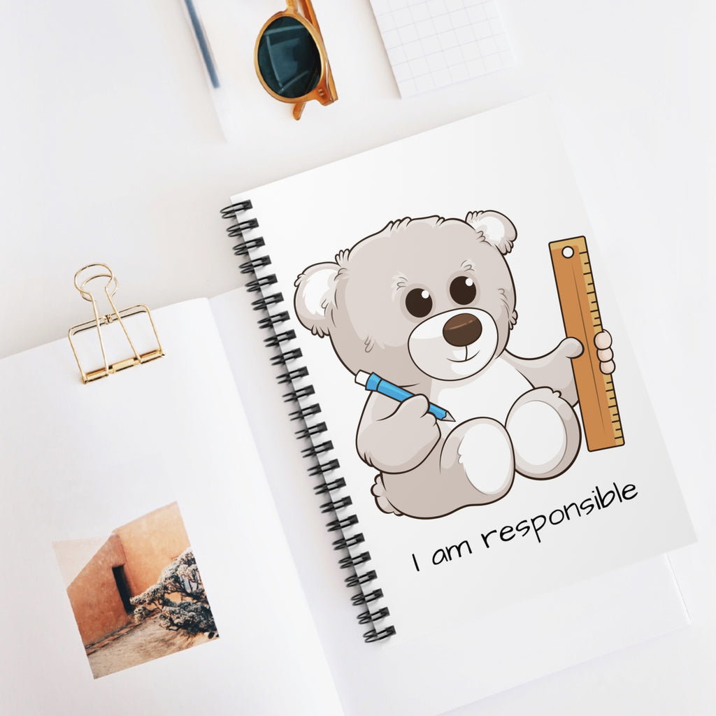 White spiral notebook with a picture of a bear that says I am responsible. The notebook is laying closed on a desk.