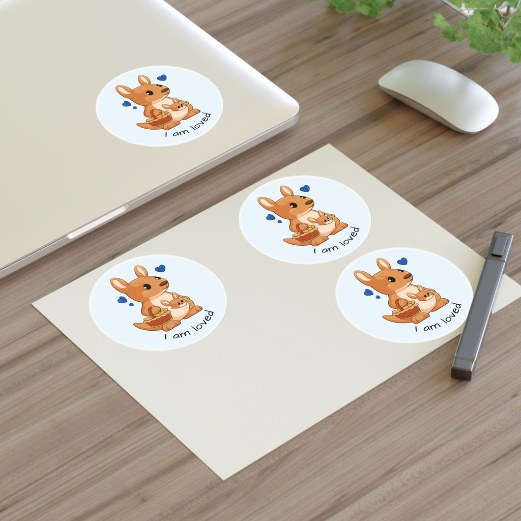 A sheet of 3 round stickers with a picture of a kangaroo that says I am loved. The sticker sheet sits on a table next to a laptop with the fourth sticker on it.