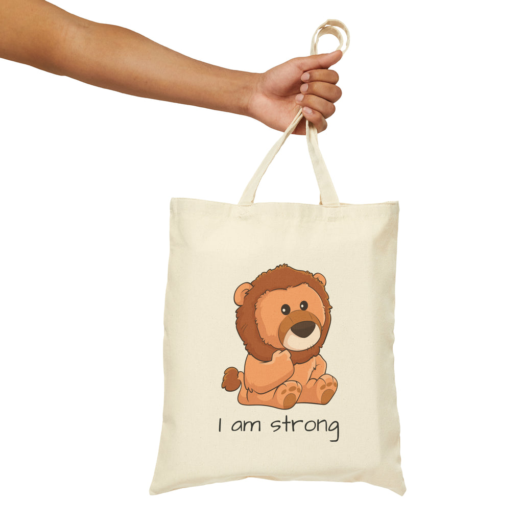 A hand holding a natural tan tote bag with a picture of a lion that says I am strong.