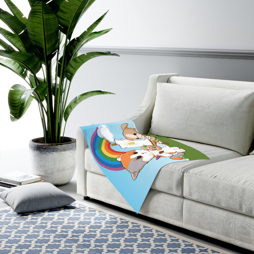Full-color swaddle blanket with a cat painting on a canvas next to a dog and a rainbow in the background. The blanket is draped over the armrest of a couch.