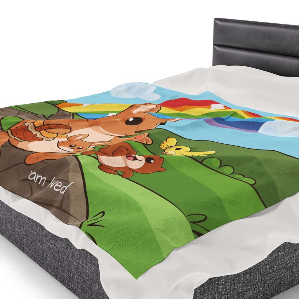 Side-view of a 60 by 80 inch blanket on a queen-sized bed. The blanket has a scene of a kangaroo walking along a path through rolling hills, a rainbow in the background, and the phrase "I am loved" along the bottom.