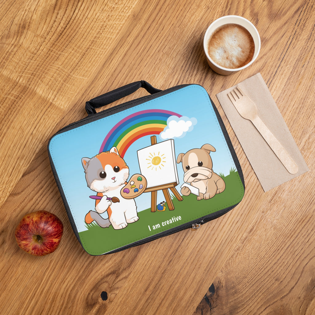 A lunch bag laying closed on a table next to a cup, fork, and apple. The lunch bag has a scene on the front of a cat painting on a canvas next to a dog, a rainbow in the background, and the phrase "I am creative" along the bottom.
