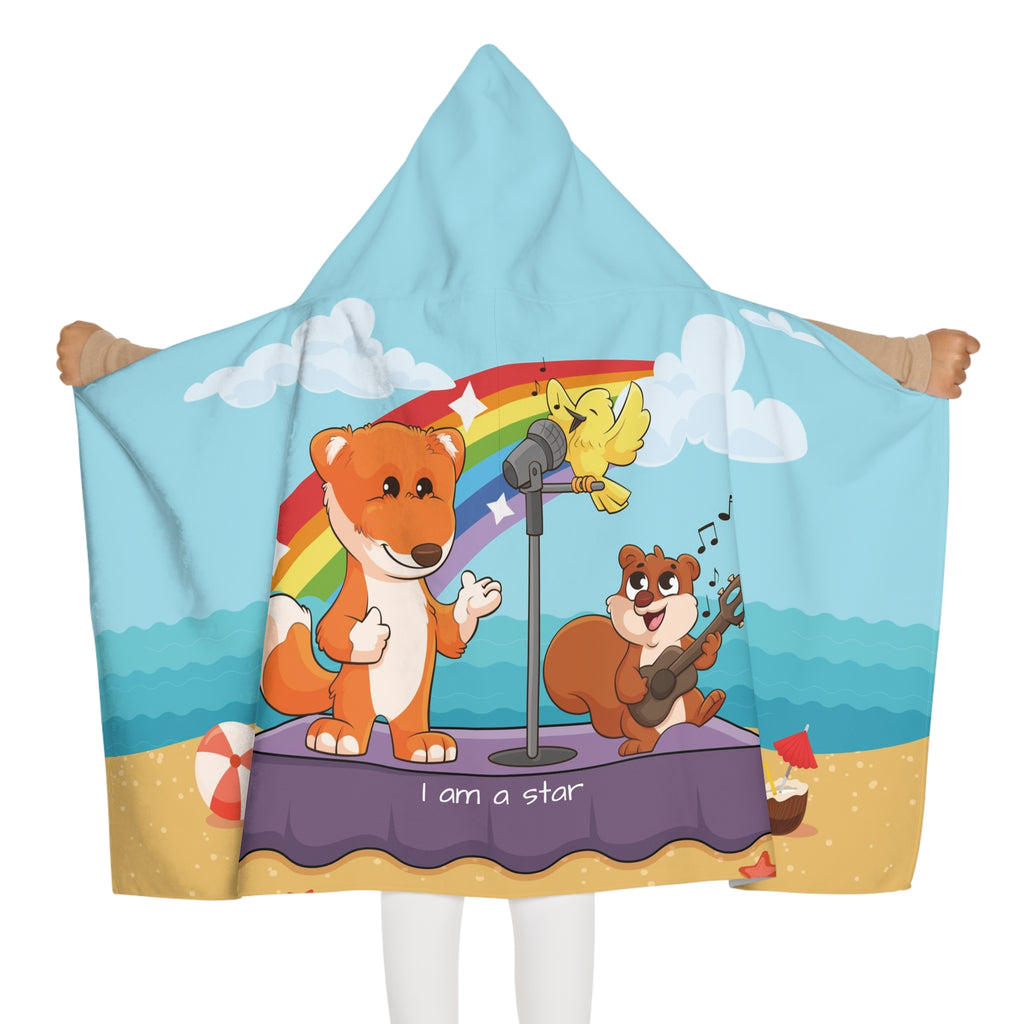 Back-view of a girl wearing a hooded towel and holding it open. The towel has a scene of a fox singing with a bird and squirrel on a stage on the beach, a rainbow in the background, and the phrase "I am a star" along the bottom.