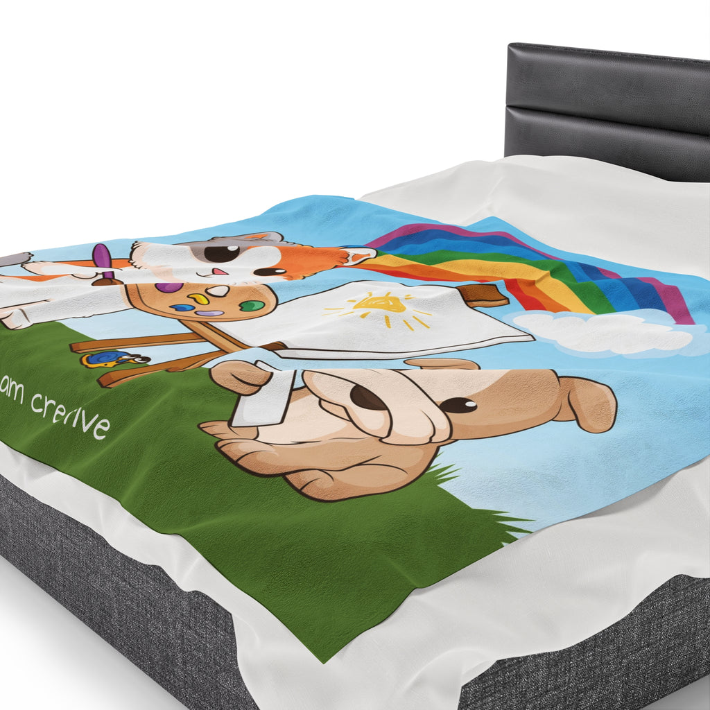 Side-view of a 60 by 80 inch blanket on a queen-sized bed. The blanket has a scene of a cat painting on a canvas next to a dog, a rainbow in the background, and the phrase "I am creative" along the bottom.