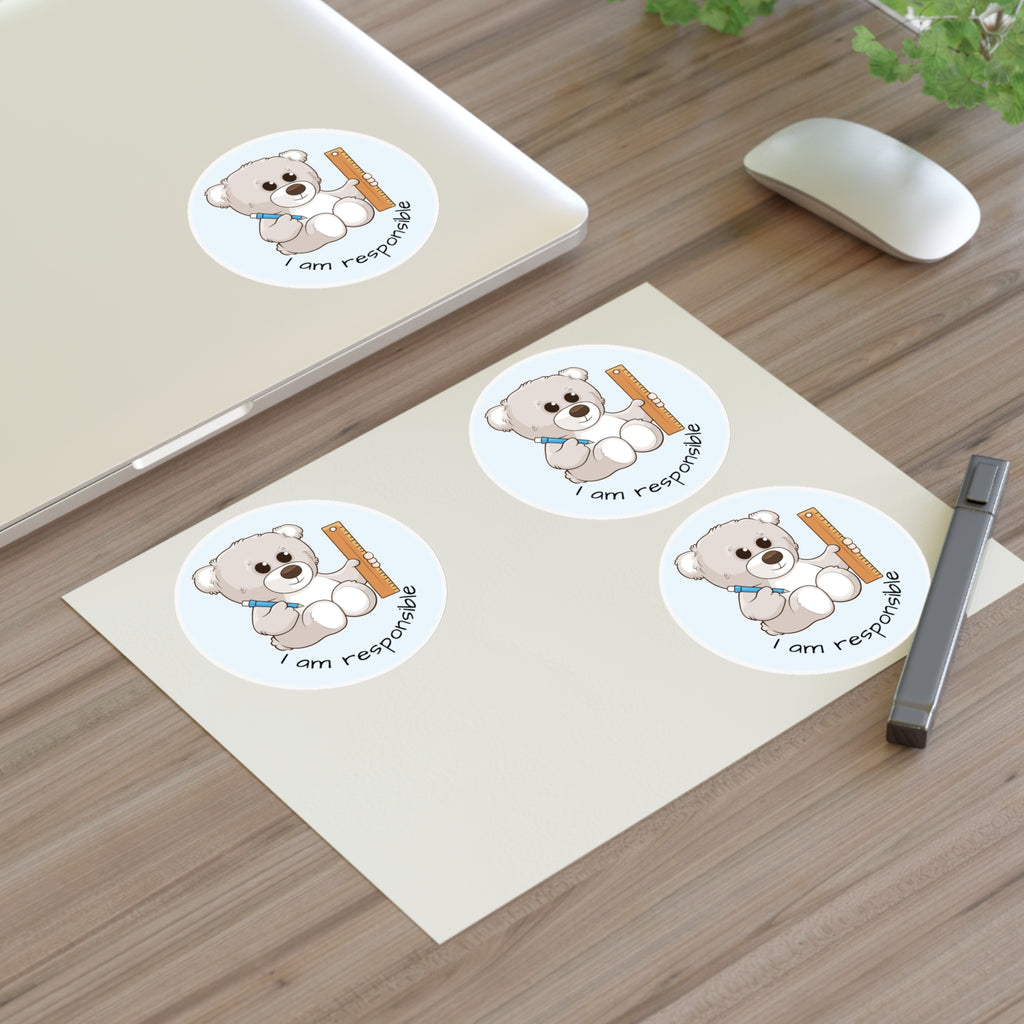 A sheet of 3 round stickers with a picture of a bear that says I am responsible. The sticker sheet sits on a table next to a laptop with the fourth sticker on it.