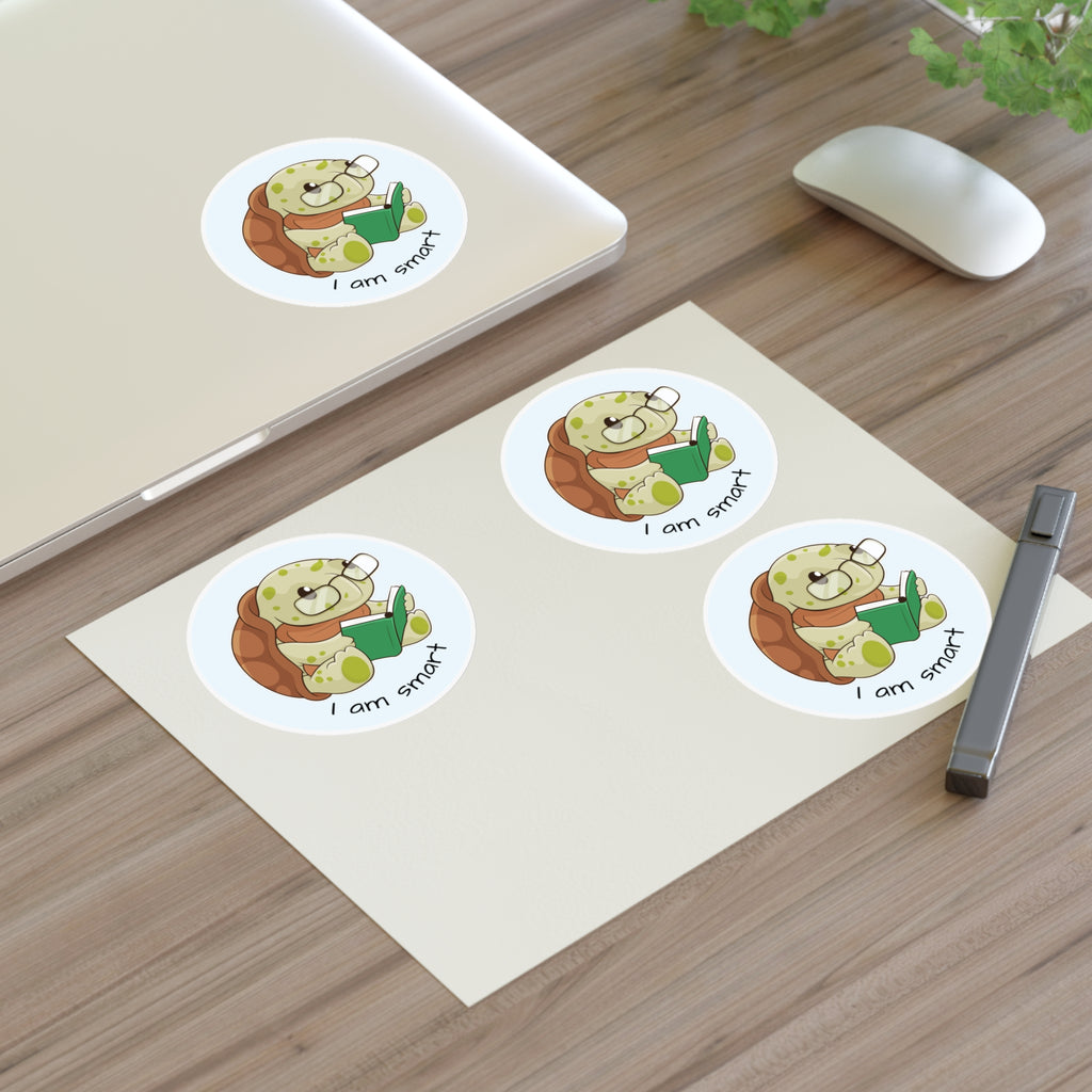 A sheet of 3 round stickers with a picture of a turtle that says I am smart. The sticker sheet sits on a table next to a laptop with the fourth sticker on it.