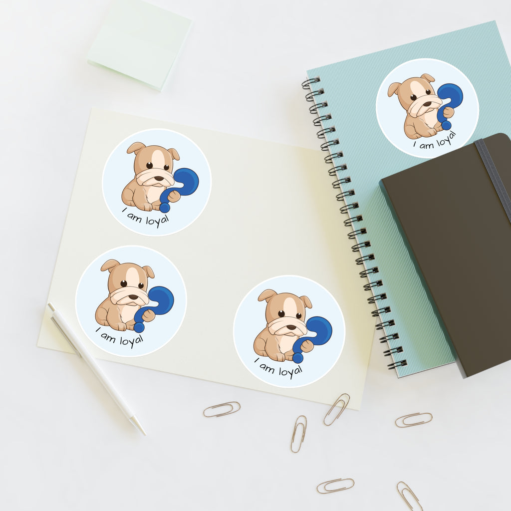 A sheet of 3 round stickers with a picture of a dog that says I am loyal. The sticker sheet sits on a table next to a notebook with the fourth sticker on it.