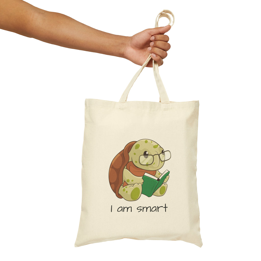 A hand holding a natural tan tote bag with a picture of a turtle that says I am smart.