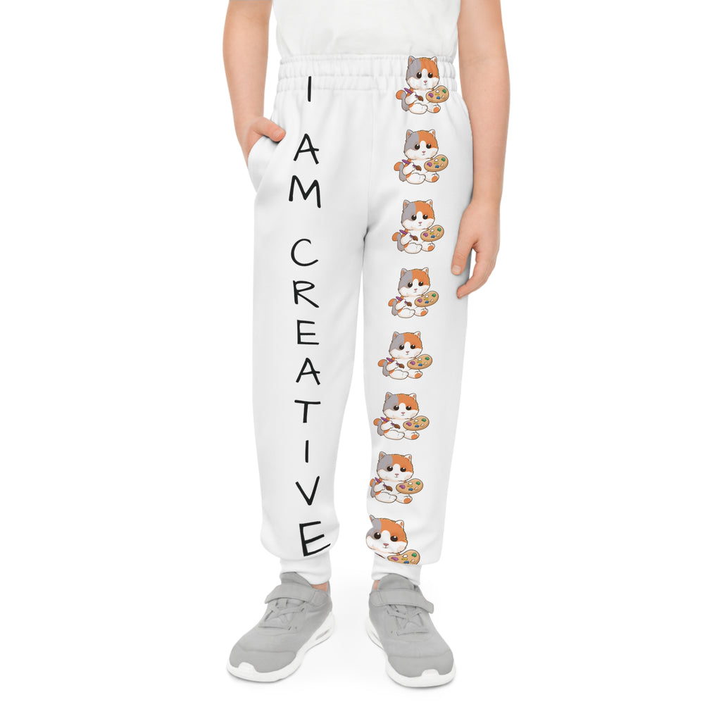 Front-view of a boy wearing white sweatpants with a line of cats down the front left leg and the phrase "I am creative" down the front right leg.