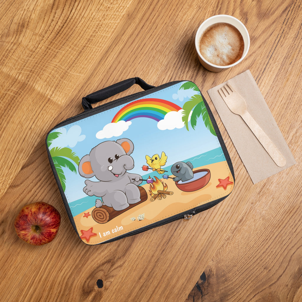 A lunch bag laying closed on a table next to a cup, fork, and apple. The lunch bag has a scene on the front of an elephant having a bonfire with a bird and fish on the beach, a rainbow in the background, and the phrase "I am calm" along the bottom.