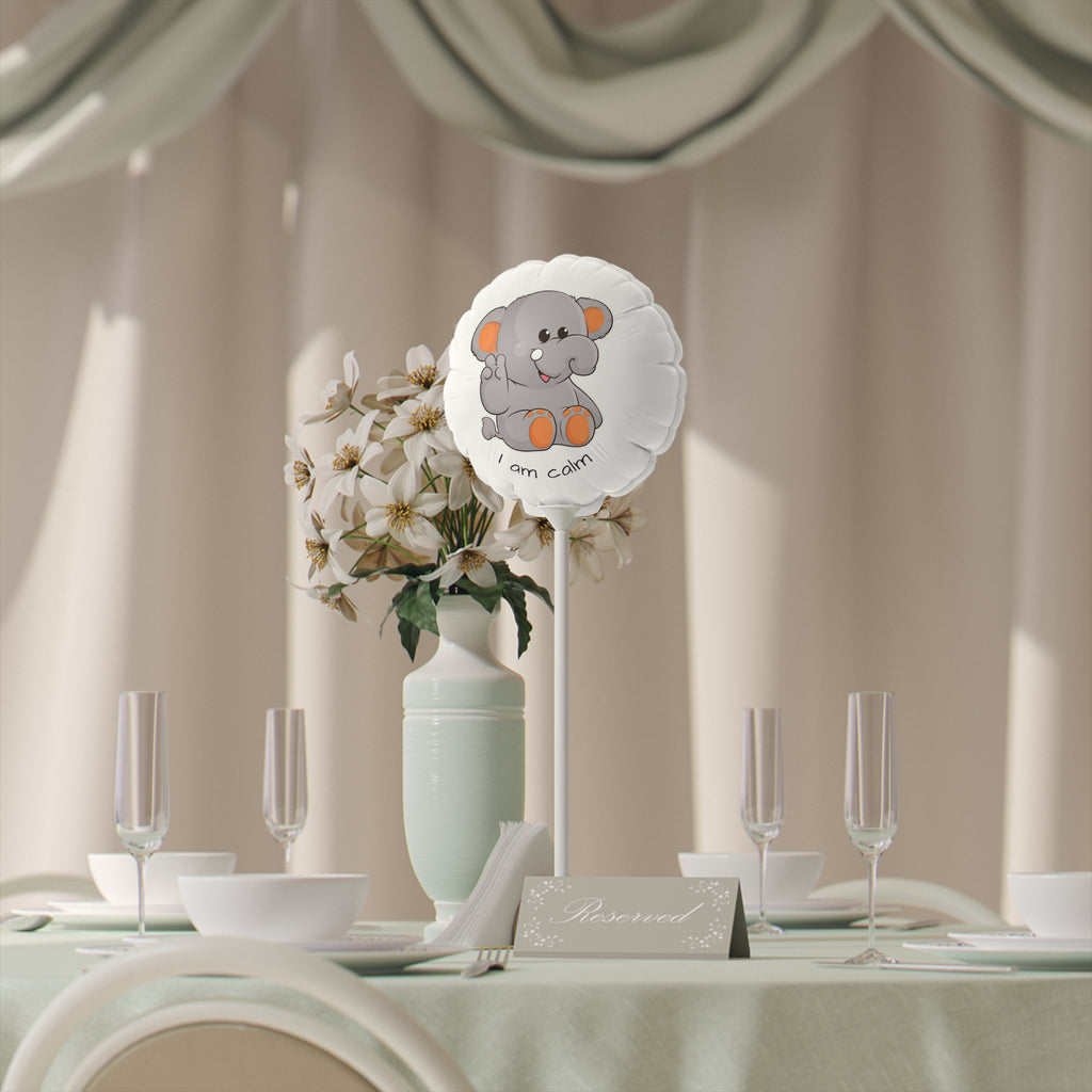 A round white mylar balloon on a stick with a picture of an elephant that says I am calm. The balloon sits on a table decorated for an event.