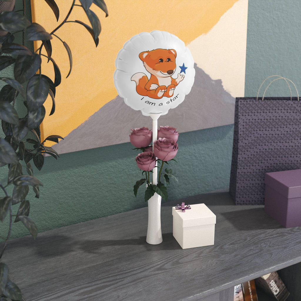 A round white mylar balloon on a stick with a picture of a fox that says I am a star. The balloon sits in a flower vase on a gift table.
