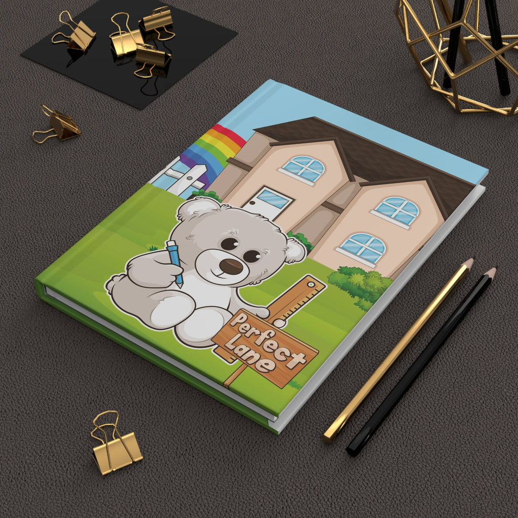 A hardcover journal laying closed on a desk. The journal cover is a scene of a bear in the yard of its house with a rainbow in the background.