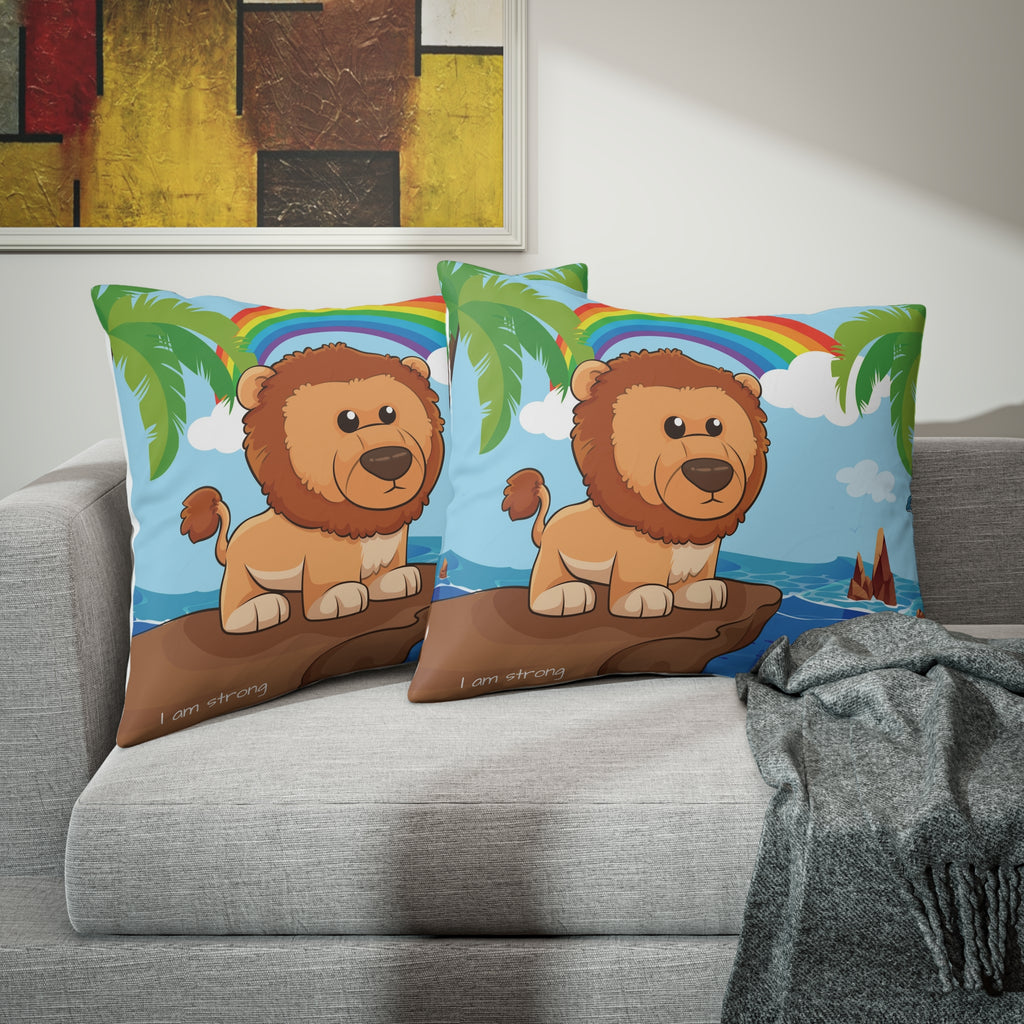 Two pillows sitting on a grey couch. The pillows have on pillowcases with a scene of a lion standing on a cliff over the ocean, a rainbow in the background, and the phrase "I am strong" along the bottom.