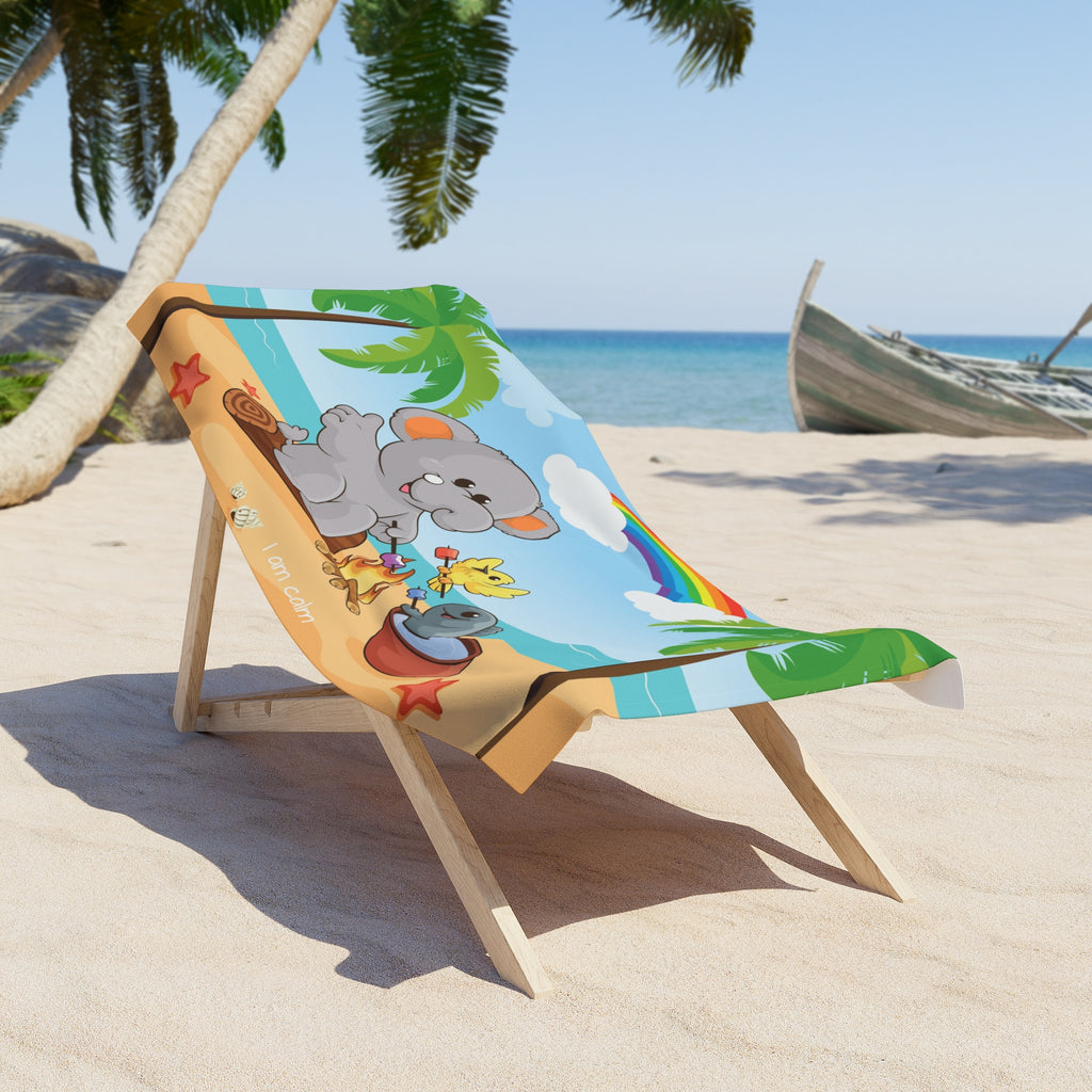A 30 by 60 inch beach towel draped over a chair on a beach. The towel has a scene of an elephant having a bonfire with a bird and fish on the beach, a rainbow in the background, and the phrase "I am calm" along the bottom.