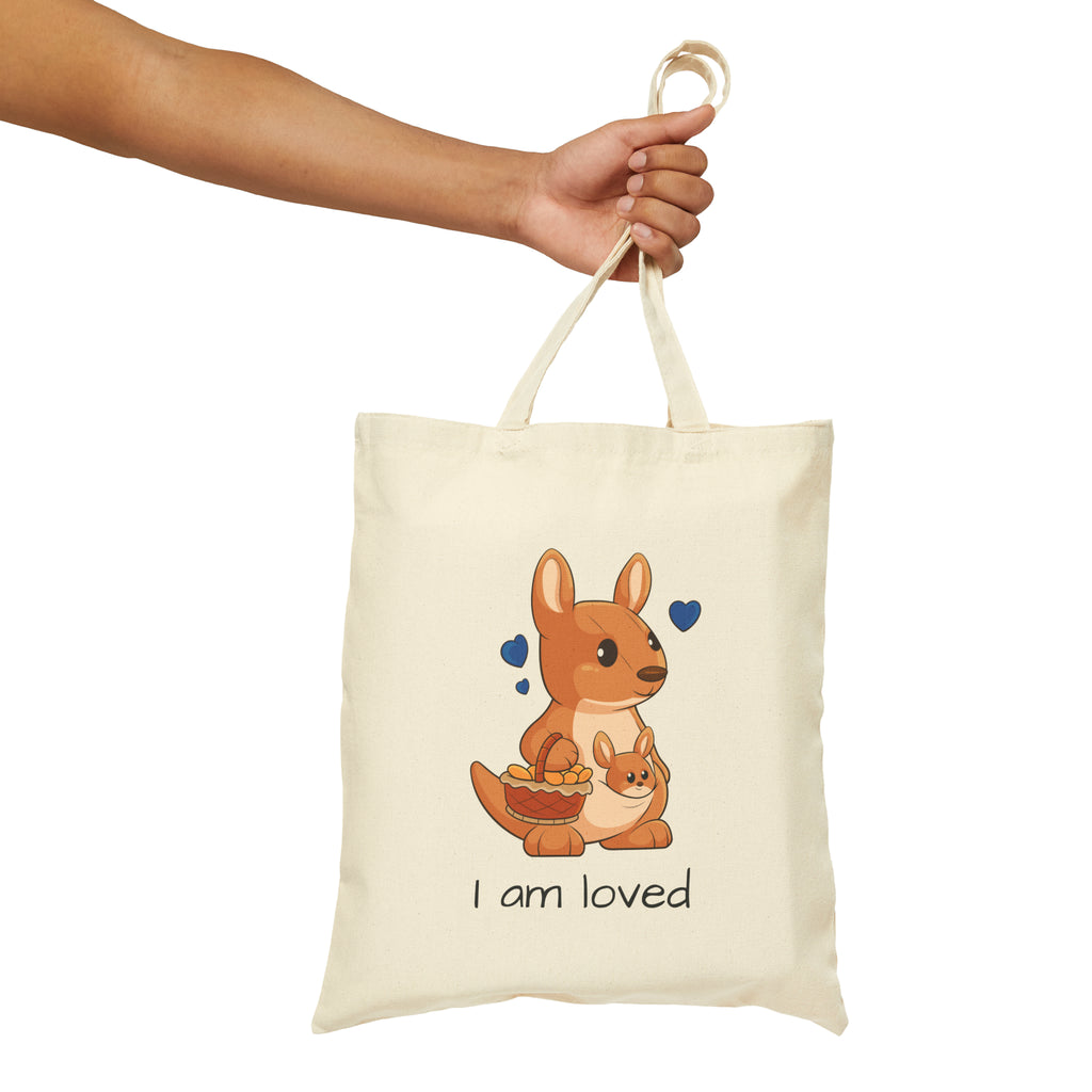 A hand holding a natural tan tote bag with a picture of a kangaroo that says I am loved.