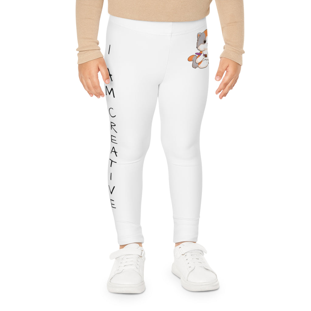 Front-view of a child wearing white leggings with a picture of a cat on the front left waist and the phrase "I am creative" read top to bottom on the side of each leg.