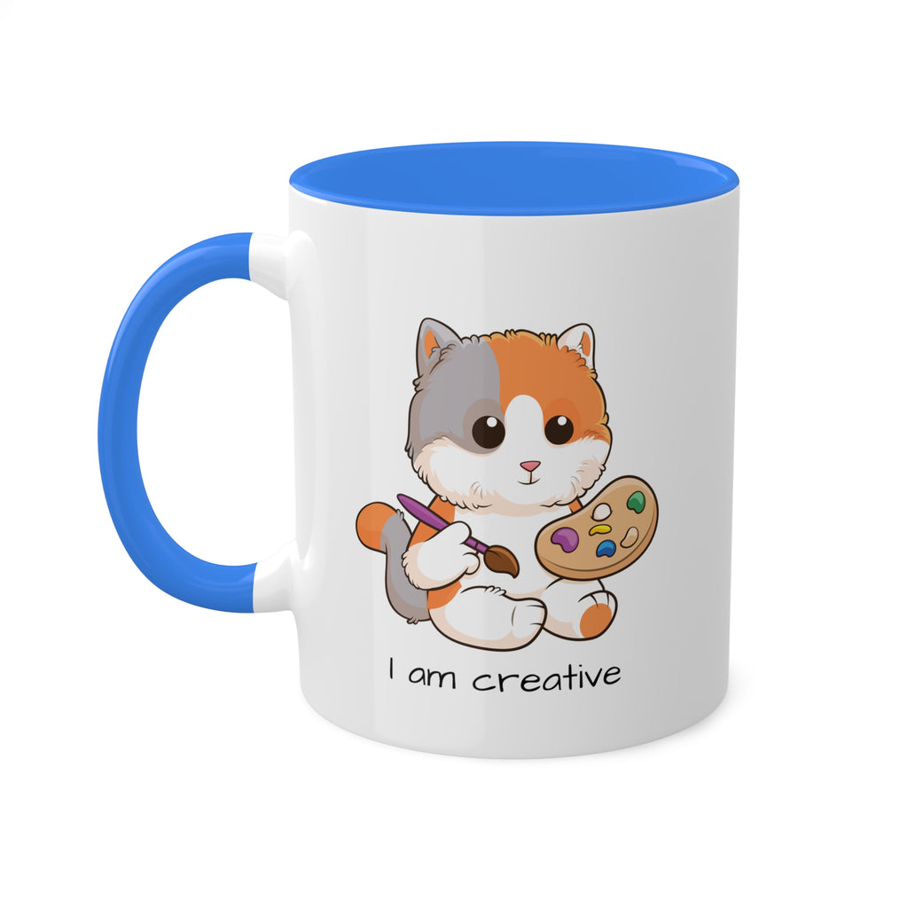A white mug with a cambridge blue handle and interior and a picture of a cat that says I am creative.