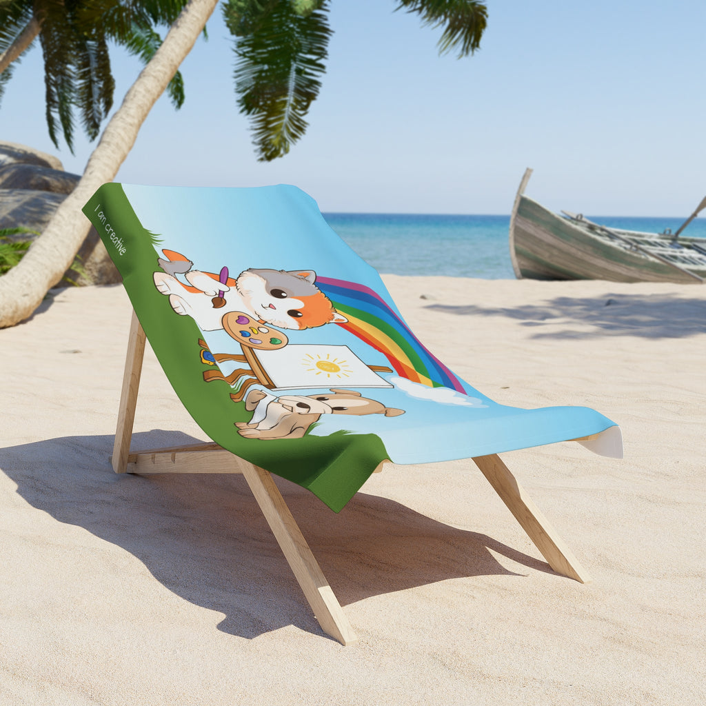 A 30 by 60 inch beach towel draped over a chair on a beach. The towel has a scene of a cat painting on a canvas next to a dog, a rainbow in the background, and the phrase "I am creative" along the bottom.