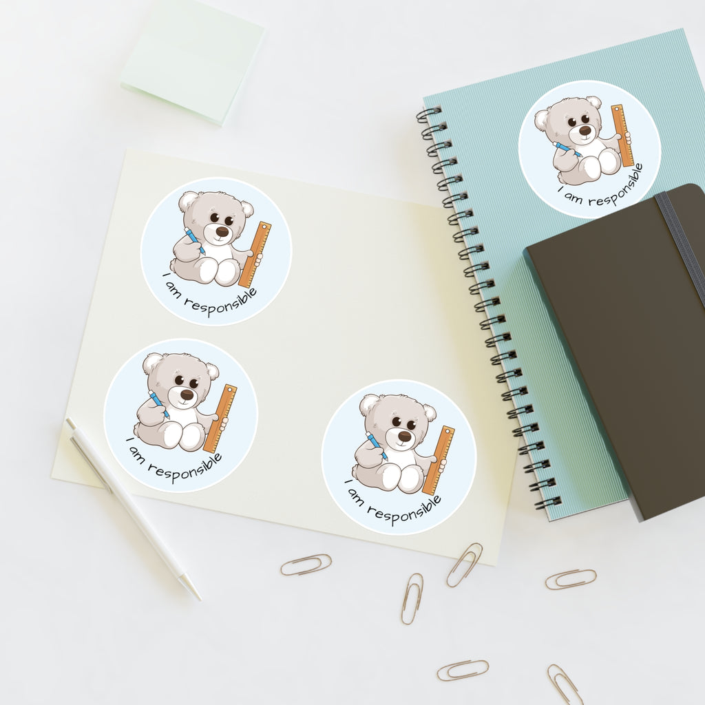 A sheet of 3 round stickers with a picture of a bear that says I am responsible. The sticker sheet sits on a table next to a notebook with the fourth sticker on it.