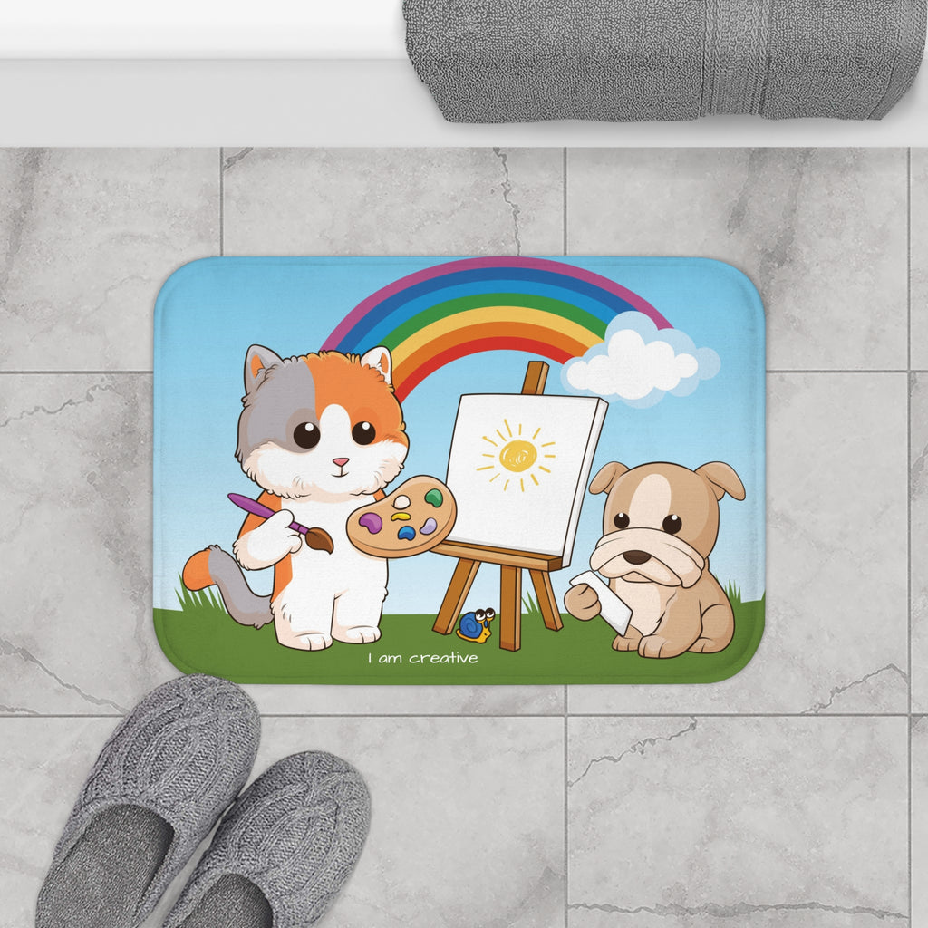 A 24 by 17 inch bath mat on the tiled floor of a bathroom. The bath mat has a scene of a cat painting on a canvas next to a dog with a rainbow in the background and the phrase "I am creative" along the bottom.