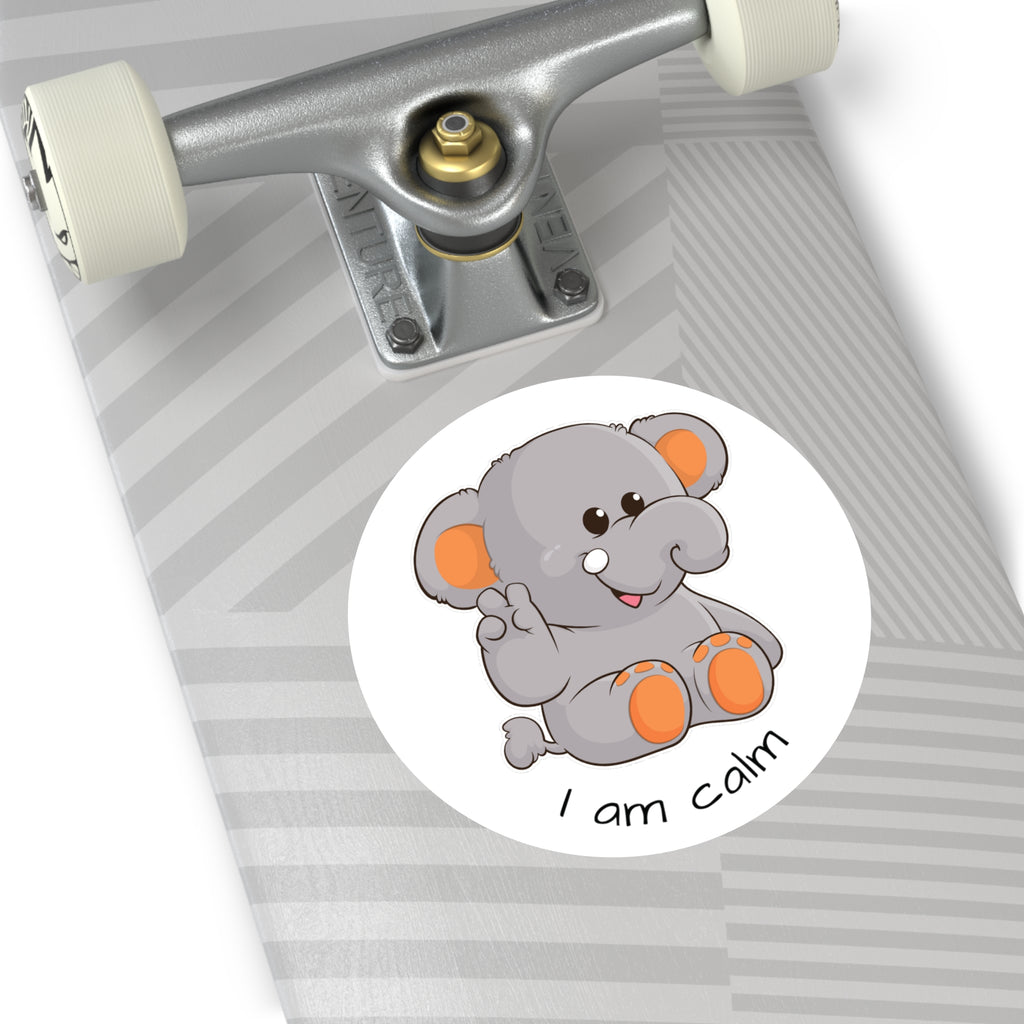 A 5 by 5 inch round white vinyl sticker with a picture of an elephant that says I am calm. The sticker is on the bottom of a grey skateboard.