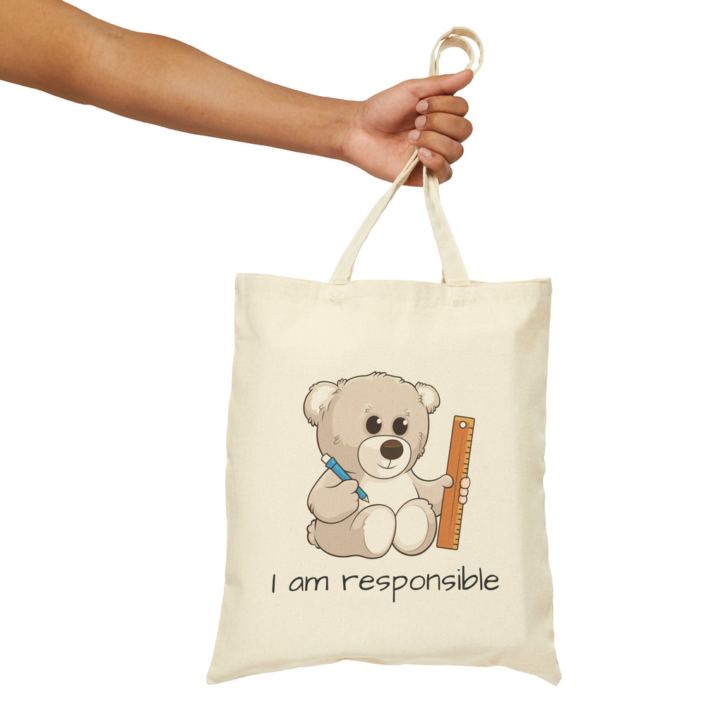 A hand holding a natural tan tote bag with a picture of a bear that says I am responsible.