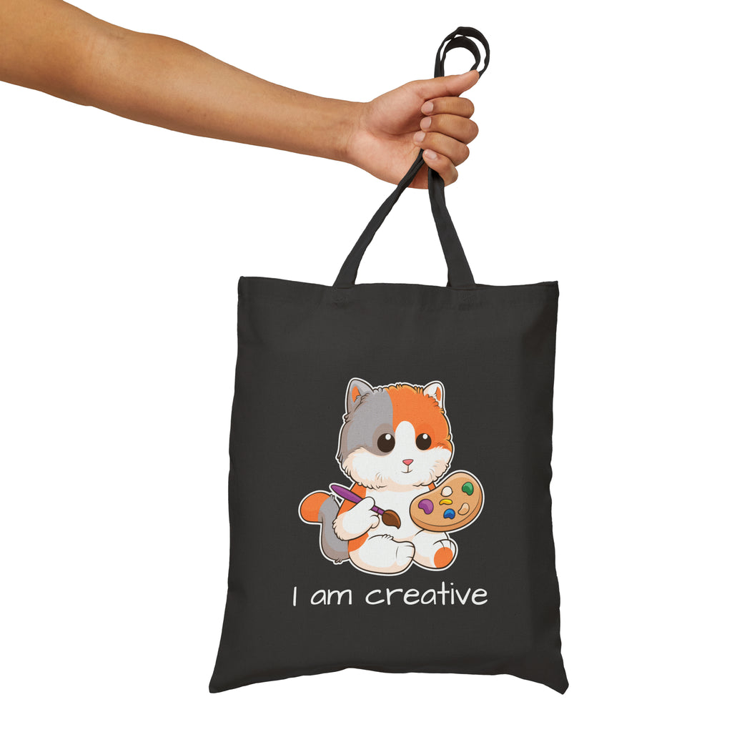 A hand holding a black tote bag with a picture of a cat that says I am creative.