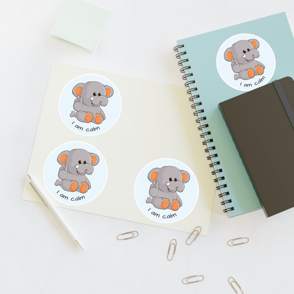 A sheet of 3 round stickers with a picture of an elephant that says I am calm. The sticker sheet sits on a table next to a notebook with the fourth sticker on it.