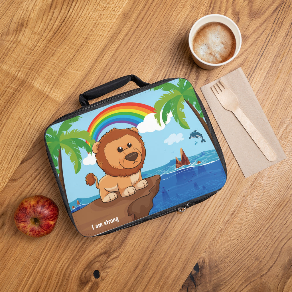 A lunch bag laying closed on a table next to a cup, fork, and apple. The lunch bag has a scene on the front of a lion standing on a cliff over the ocean, a rainbow in the background, and the phrase "I am strong" along the bottom.
