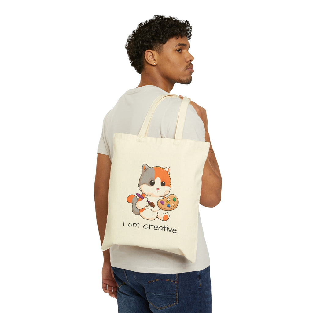 A man with a natural tan tote bag over his shoulder, featuring a picture of a cat that says I am creative.
