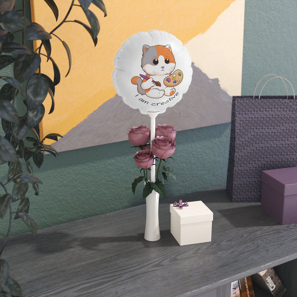 A round white mylar balloon on a stick with a picture of a cat that says I am creative. The balloon sits in a flower vase on a gift table.