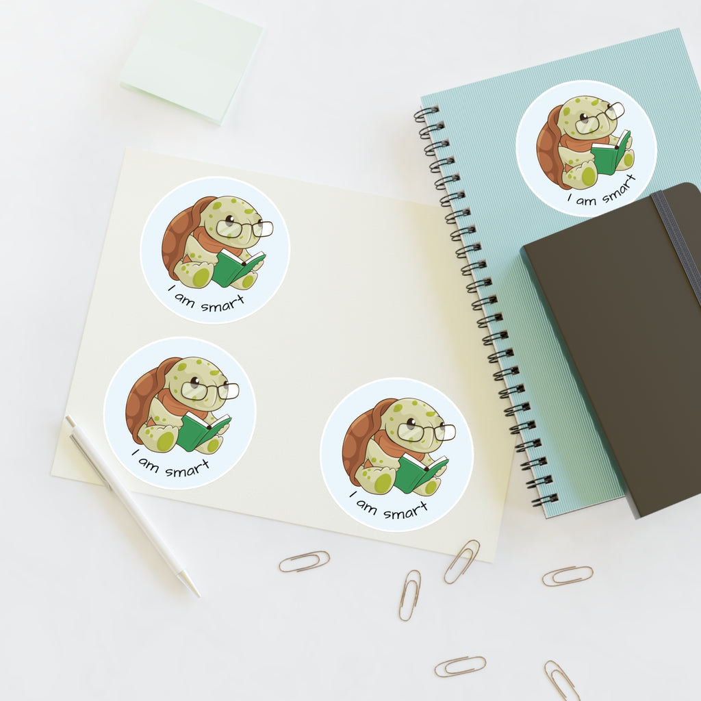 A sheet of 3 round stickers with a picture of a turtle that says I am smart. The sticker sheet sits on a table next to a notebook with the fourth sticker on it.