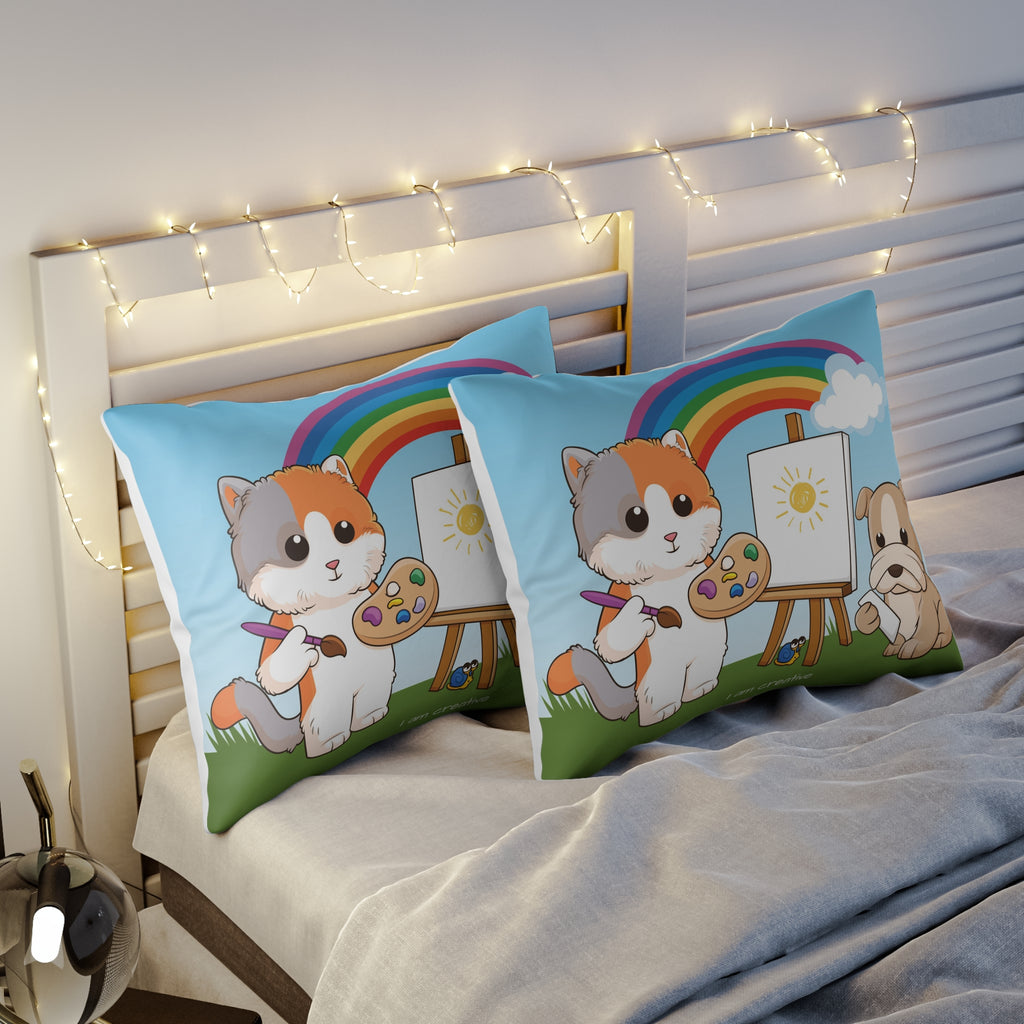 Two pillows sitting on a bed. The pillows have on pillowcases with a scene of a cat painting on a canvas next to a dog, a rainbow in the background, and the phrase "I am creative" along the bottom.