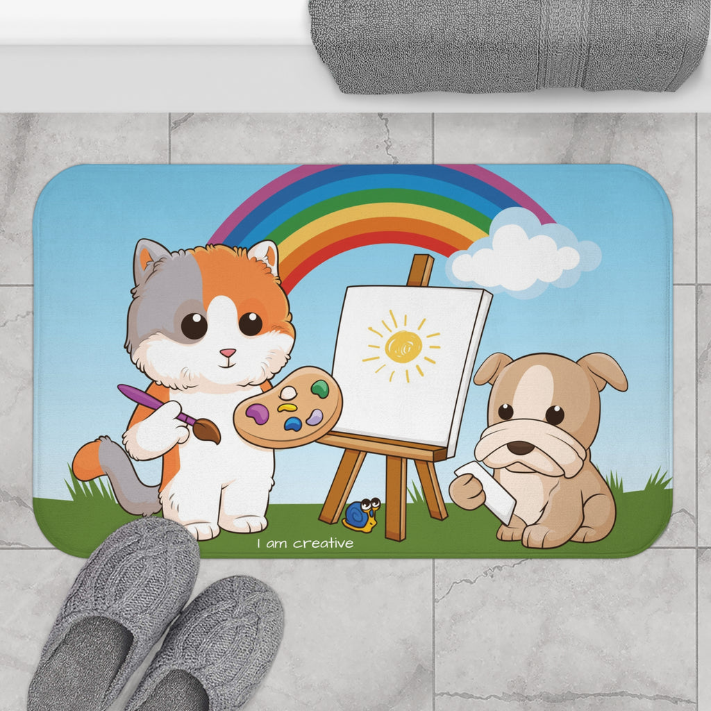 A 34 by 21 inch bath mat on the tiled floor of a bathroom. The bath mat has a scene of a cat painting on a canvas next to a dog with a rainbow in the background and the phrase "I am creative" along the bottom.