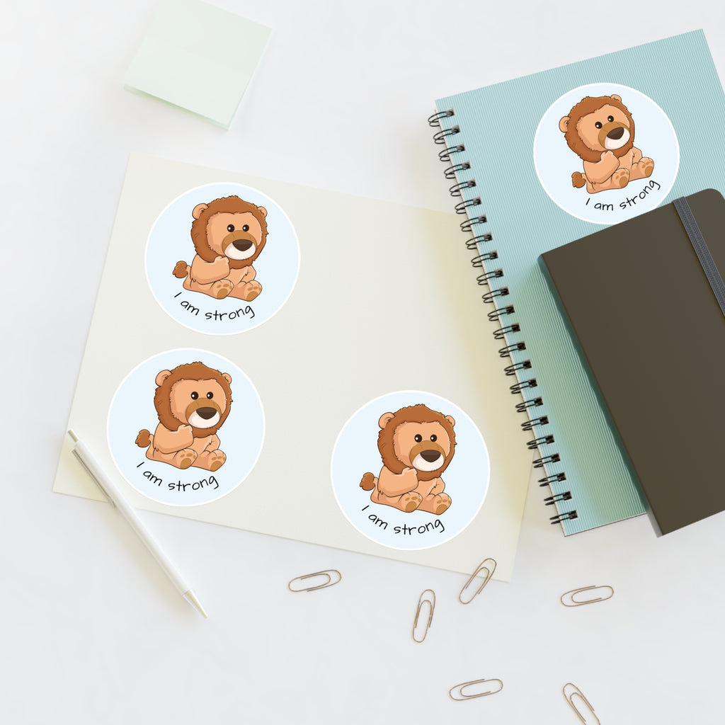 A sheet of 3 round stickers with a picture of a lion that says I am strong. The sticker sheet sits on a table next to a notebook with the fourth sticker on it.