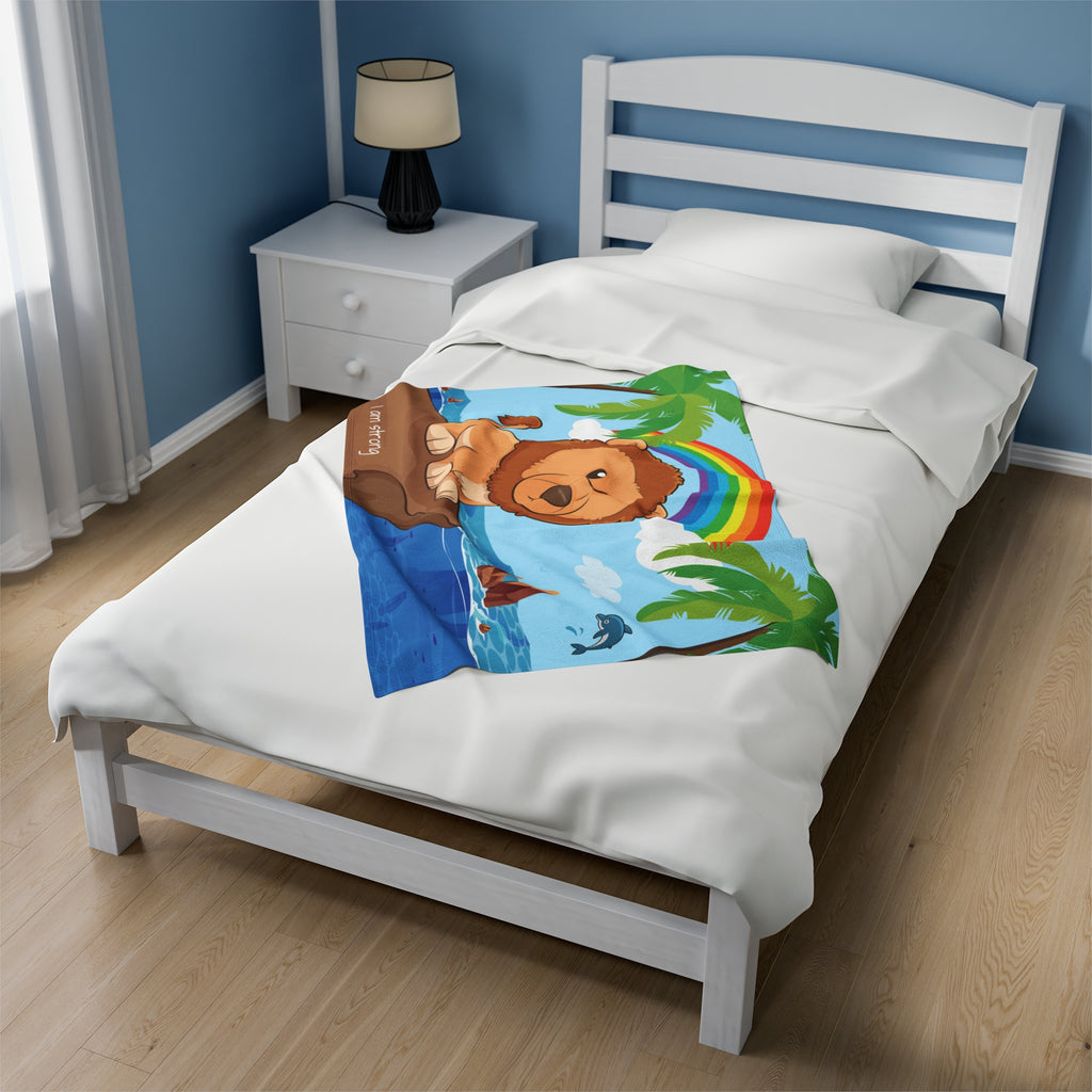 A 30 by 40 inch blanket on a twin-sized bed in a bedroom. The blanket has a scene of a lion standing on a cliff over the ocean, a rainbow in the background, and the phrase "I am strong" along the bottom.