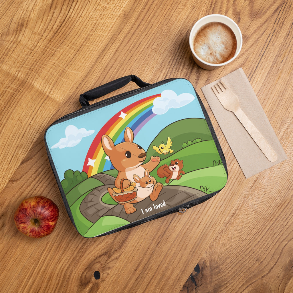 A lunch bag laying closed on a table next to a cup, fork, and apple. The lunch bag has a scene on the front of a kangaroo walking along a path through rolling hills, a rainbow in the background, and the phrase "I am loved" along the bottom.