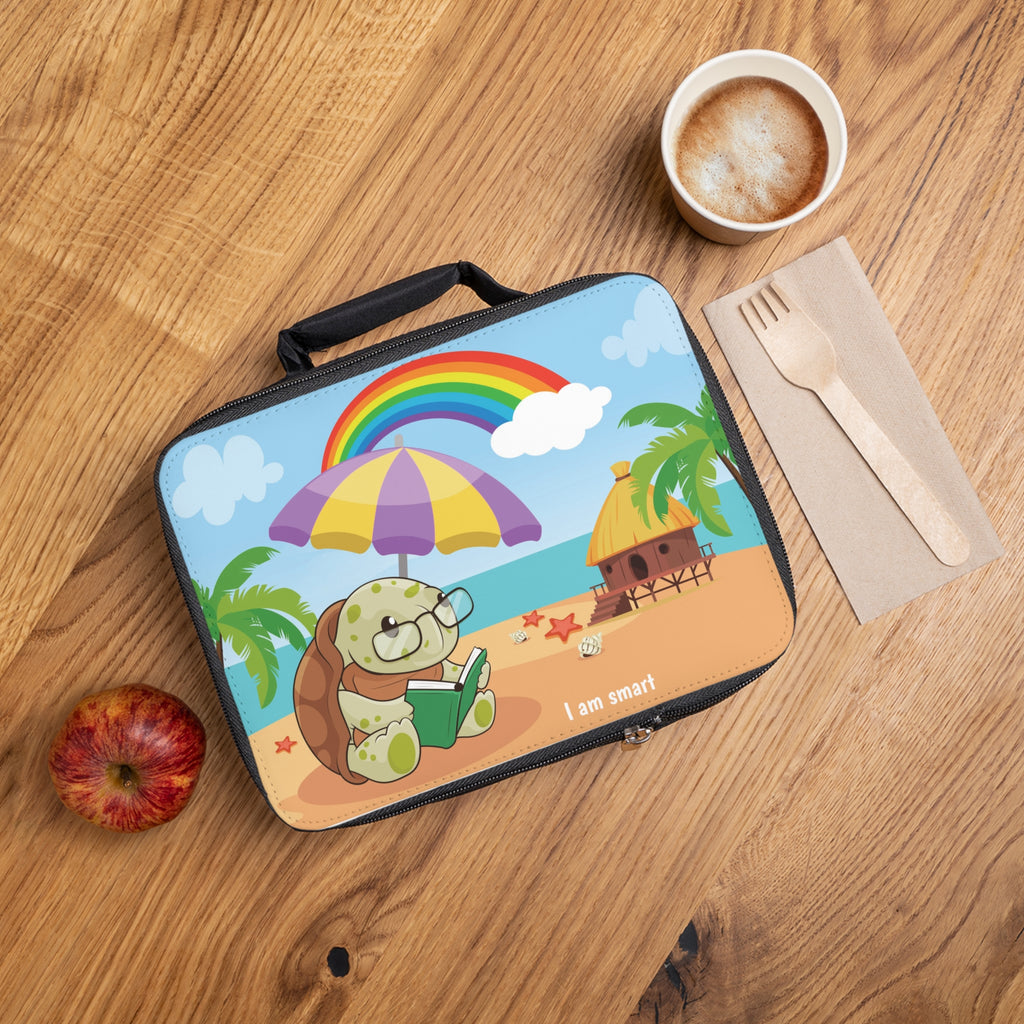A lunch bag laying closed on a table next to a cup, fork, and apple. The lunch bag has a scene on the front of a turtle reading a book under an umbrella on the beach, a rainbow in the background, and the phrase "I am smart" along the bottom.