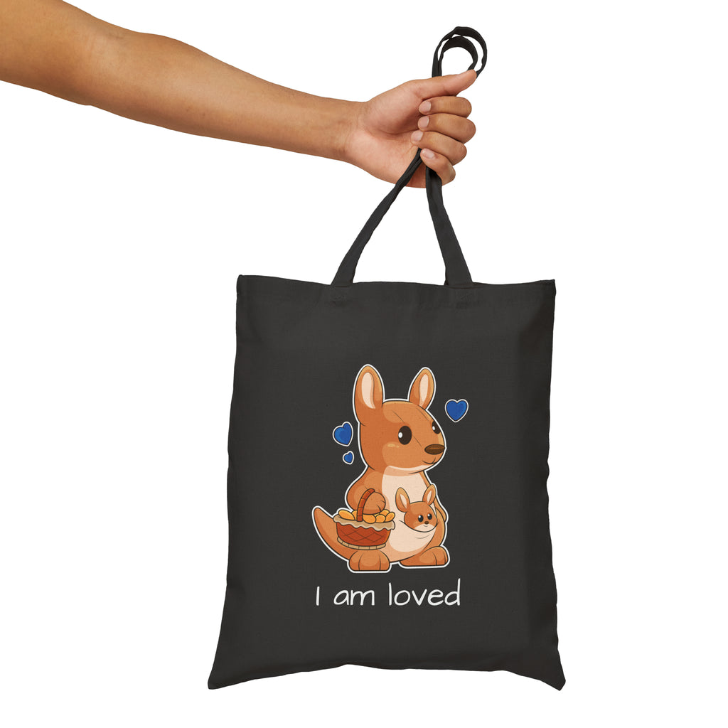 A hand holding a black tote bag with a picture of a kangaroo that says I am loved.